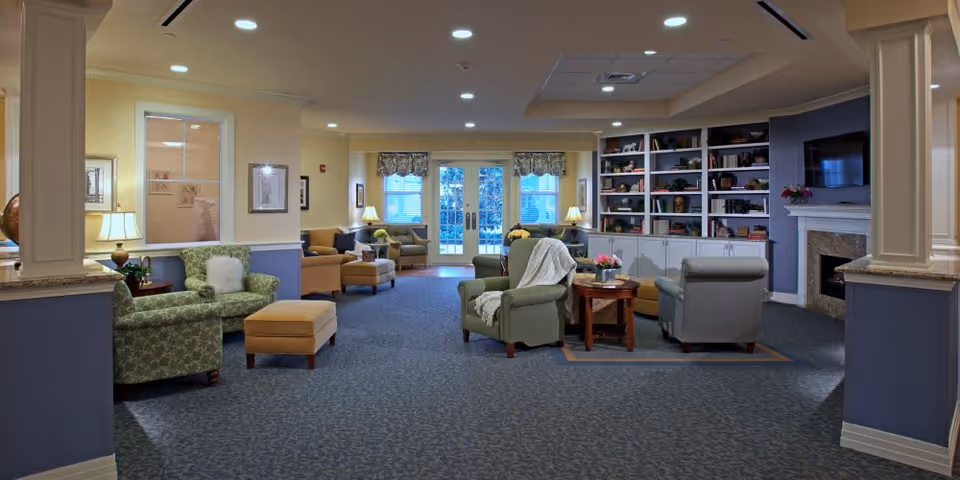 A spacious and well-lit living room area in a senior living facility featuring multiple comfortable armchairs and ottomans arranged around small tables. The room has a blue carpet, cream and blue walls, built-in bookshelves filled with decorative items, a fireplace with a mounted TV above it, and large windows with floral valances letting in natural light.