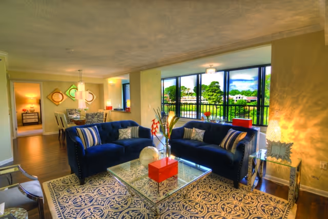A bright and spacious living room with two dark blue sofas facing each other across a glass coffee table on a patterned rug. The room has large windows letting in natural light and offering a view of greenery outside. In the background, there is a dining area with a table and chairs, and a glimpse of a bedroom through an open doorway.