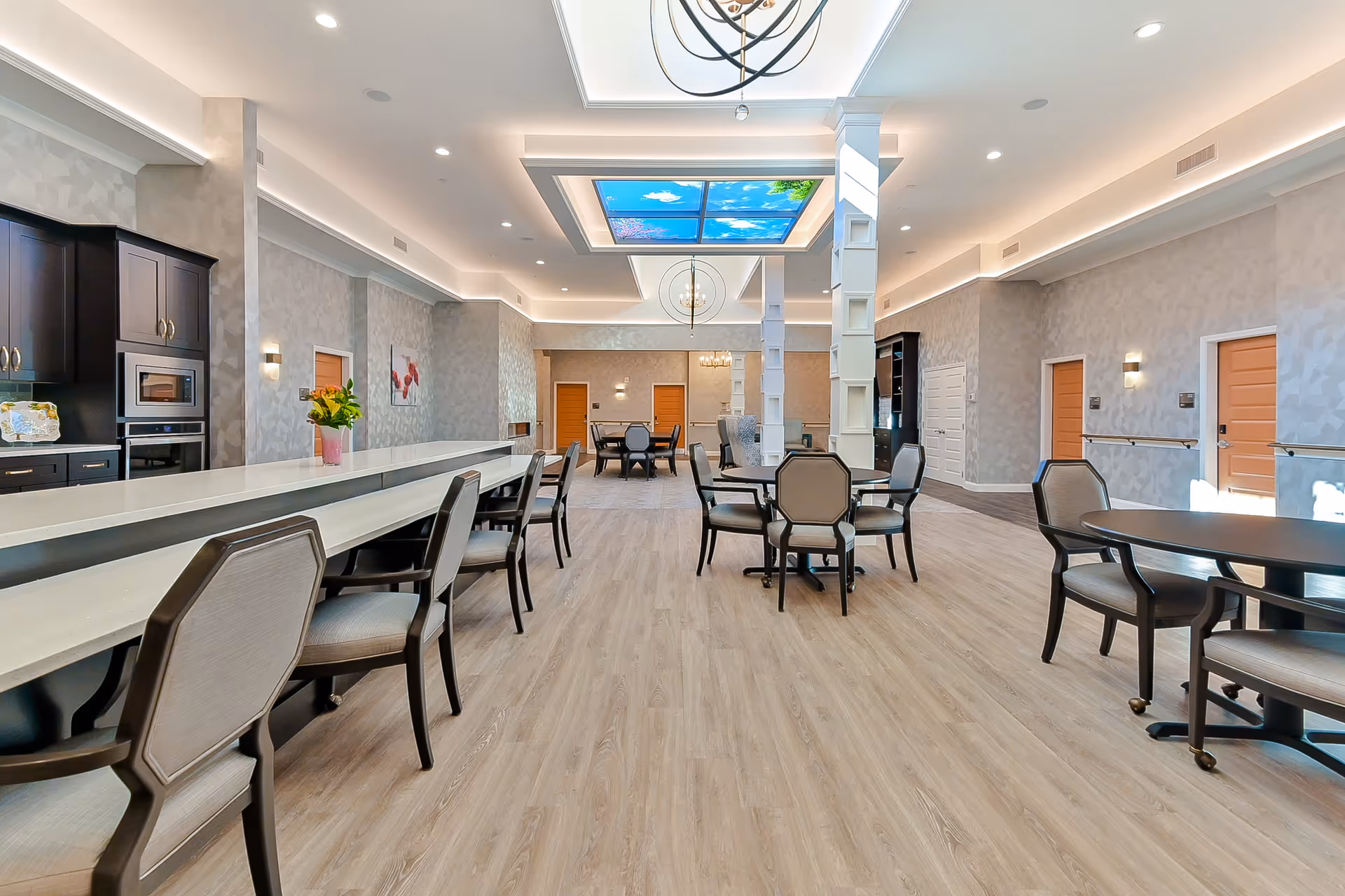 A spacious, well-lit common area with multiple seating arrangements including tables and chairs. The room features light wood flooring, modern light fixtures, and a ceiling panel displaying a sky scene with clouds and tree branches. There are cabinets and a countertop on the left side, and several doors along the walls.