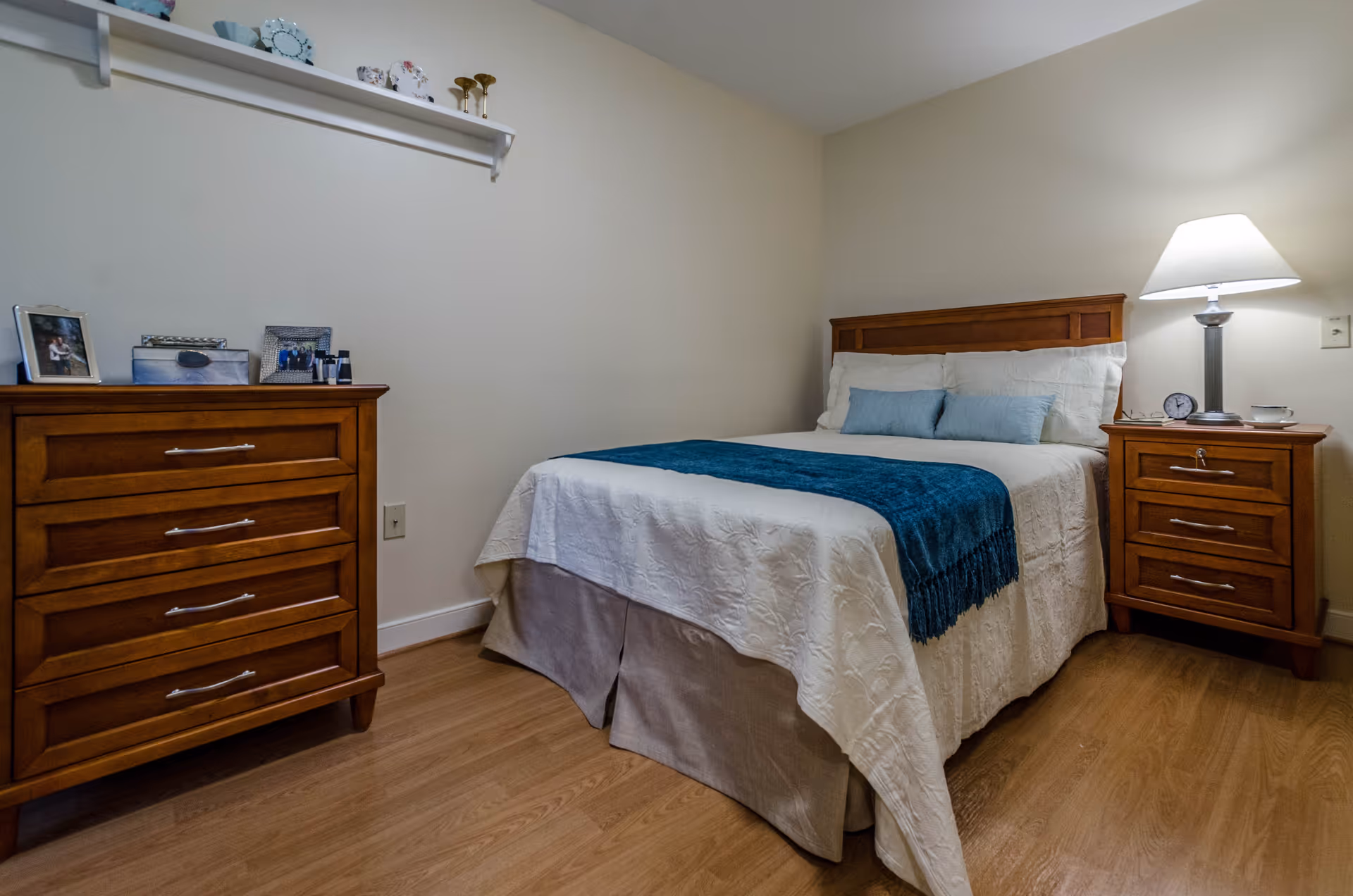 A cozy bedroom with a wooden bed frame, white bedding, and a blue throw blanket. There is a wooden nightstand with a lamp, clock, and cup on the right side of the bed. On the left side of the room, there is a wooden dresser with framed photos and decorative items on top. A white shelf with small decorative items is mounted on the wall above the dresser. The room has light-colored walls and wood flooring.