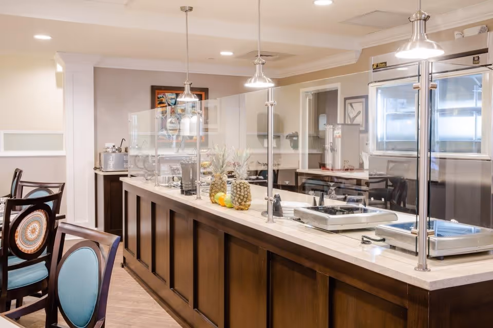 Interior view of a dining area with a serving counter featuring a glass sneeze guard, two pineapples, and various kitchen equipment. There are wooden chairs with blue cushions and decorative backs, pendant lights hanging from the ceiling, and framed artwork on the walls.