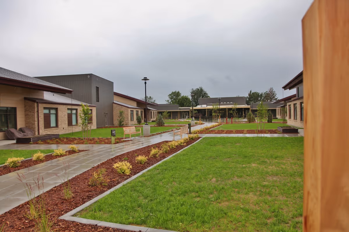 Outdoor courtyard area of Walla Walla Veterans Home featuring paved walkways, green lawns, small trees, benches, and surrounding single-story buildings under a cloudy sky.