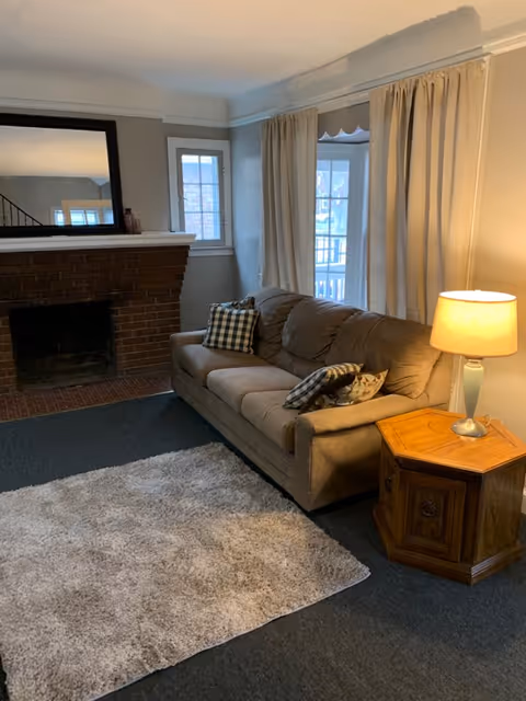 A cozy living room with a brown sofa adorned with patterned pillows, a wooden side table with a lit lamp, a large mirror above a brick fireplace, and a light-colored rug on a dark carpeted floor. There are windows with beige curtains allowing natural light into the room.