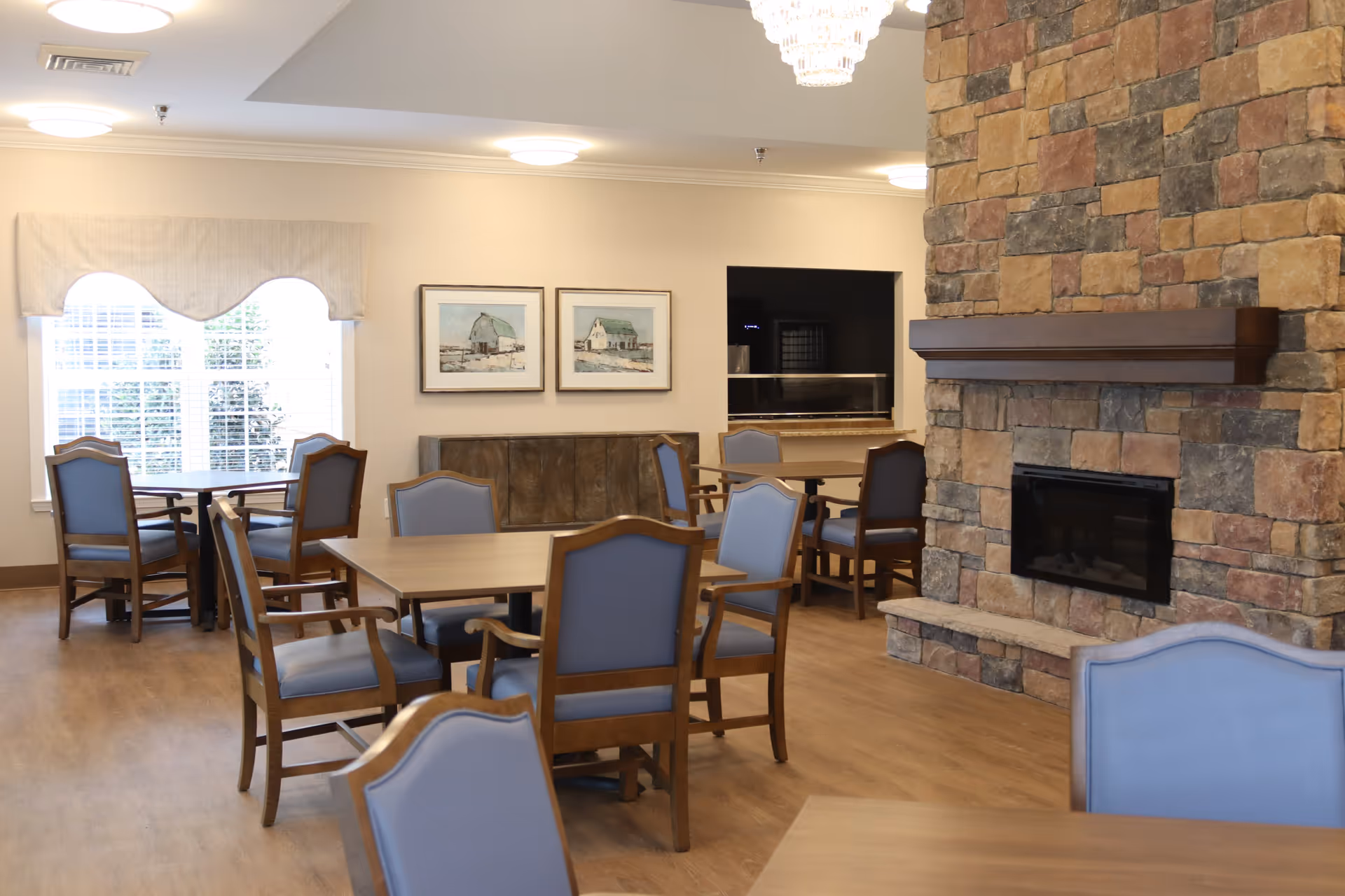 A cozy dining area with wooden tables and chairs featuring blue cushions. The room has a large stone fireplace on the right side and two framed pictures on the wall. There is a window with white blinds and a beige valance letting in natural light. The floor is wooden, and the ceiling has round light fixtures.