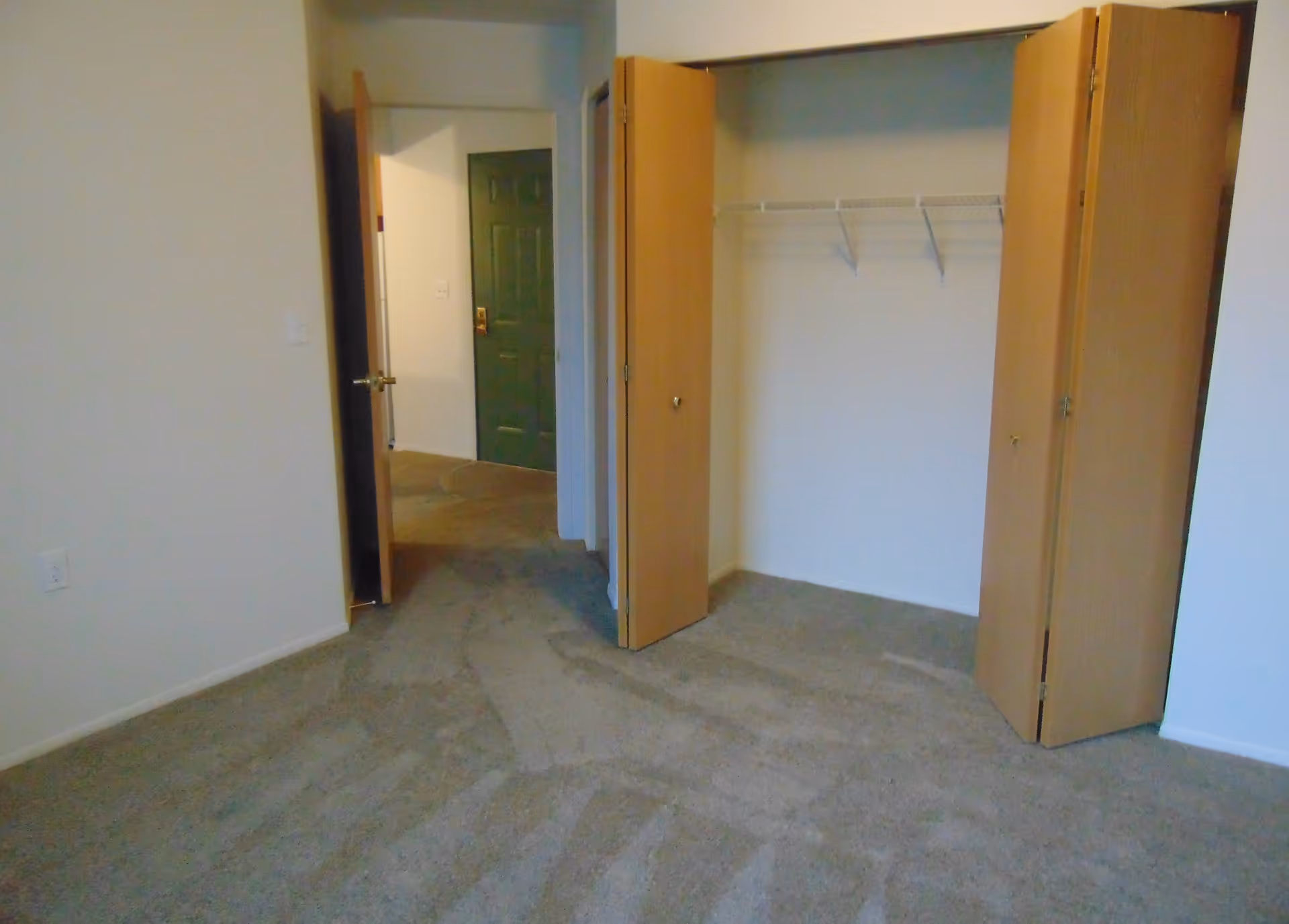 Empty room with beige carpet, white walls, an open closet with wooden bi-fold doors, and an open door leading to a hallway with a green door at the end.