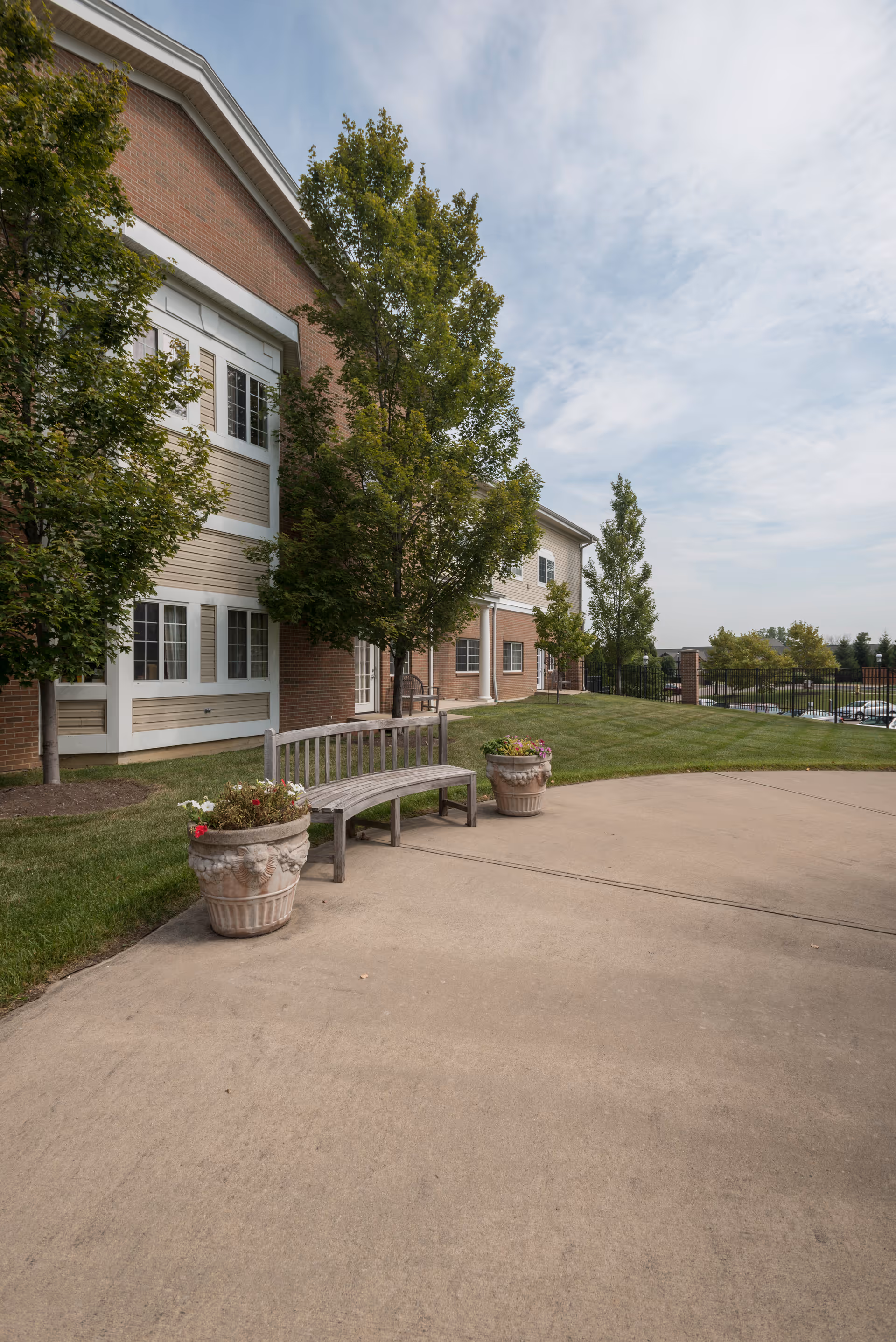 Exterior view of a brick senior living building with a wooden bench between two planters on a paved walkway and trees on a grassy lawn.