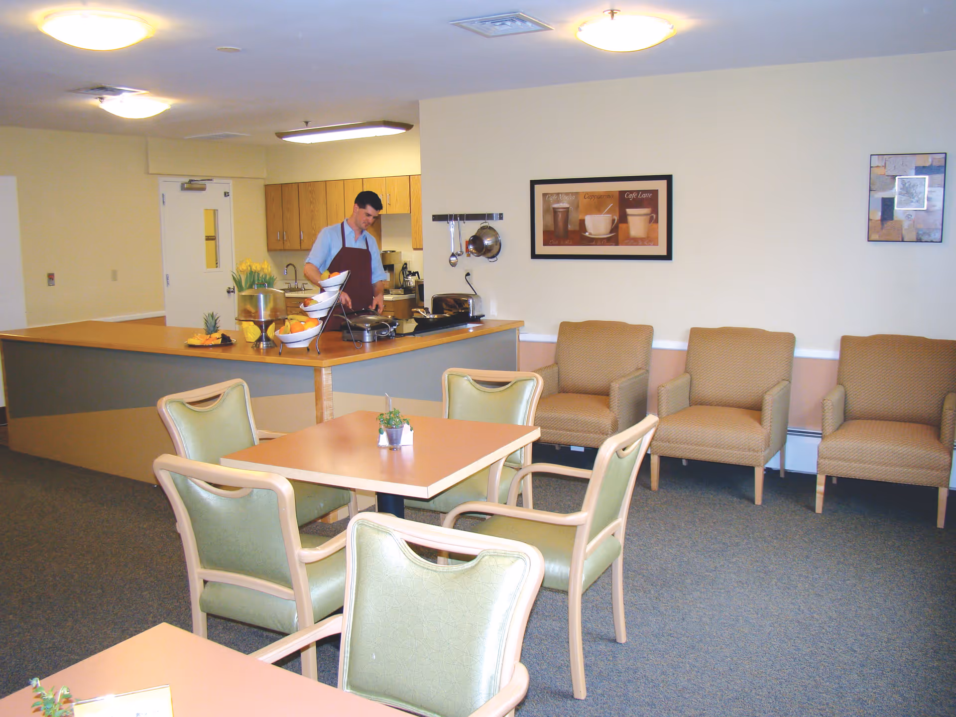 Dining/common room with tables and green chairs, a service counter where a staff member prepares food, and armchairs along the wall.