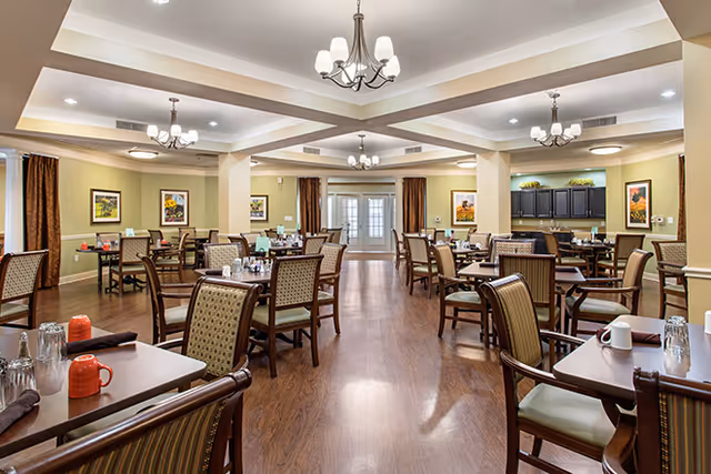 A spacious dining room with multiple wooden tables and cushioned chairs arranged neatly. Each table is set with glasses, cups, and napkins. The room features warm lighting from ceiling chandeliers, framed artwork on the walls, and large windows with brown curtains. There is a cabinet with decorative items in the background.