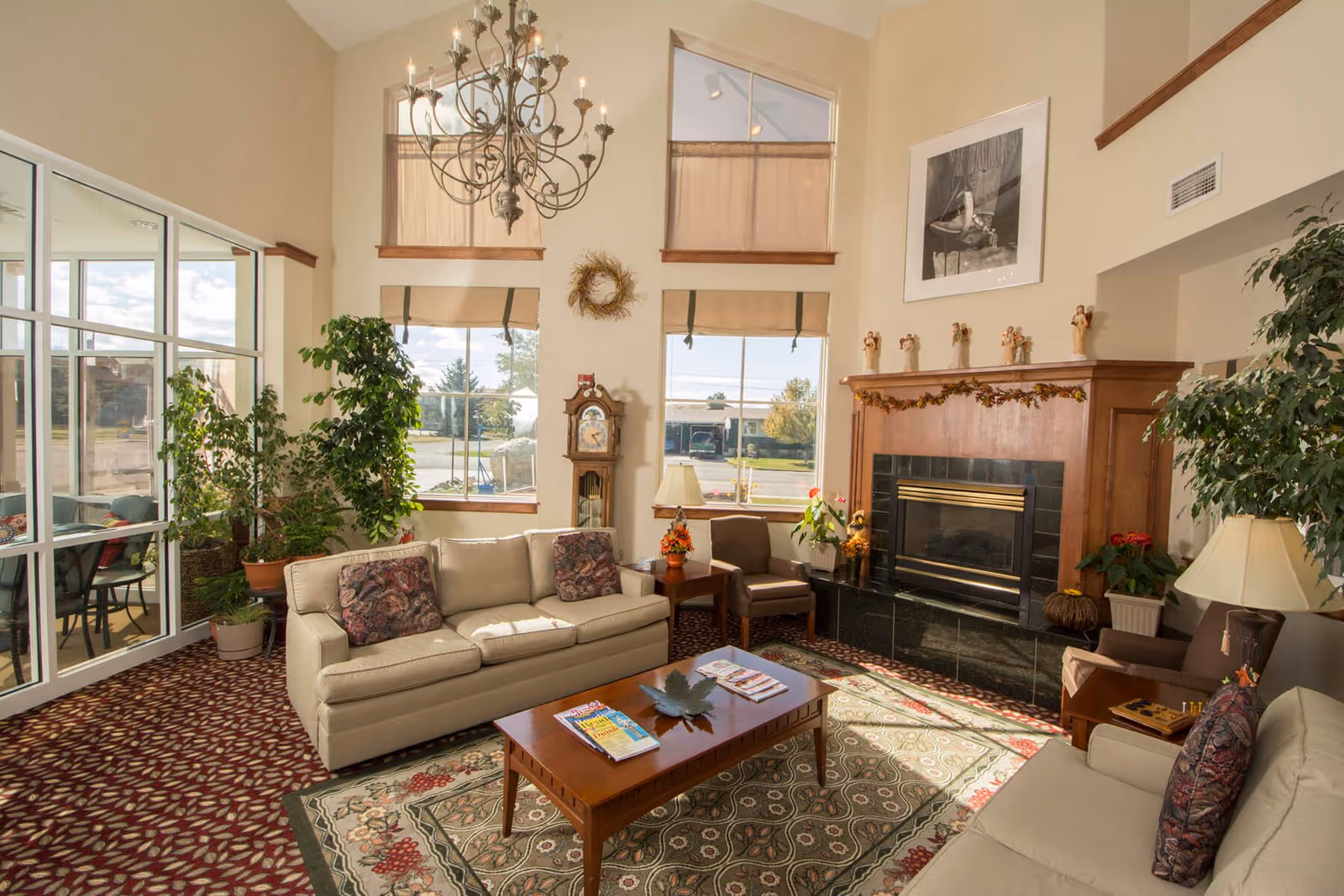 Sunlit assisted-living lounge with sofas, a coffee table, fireplace, tall windows, plants and a chandelier.