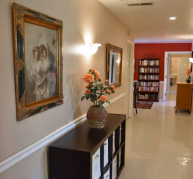 Hallway interior with a console table topped by a vase of flowers, framed artwork on the wall, and a bookshelf visible down the corridor.