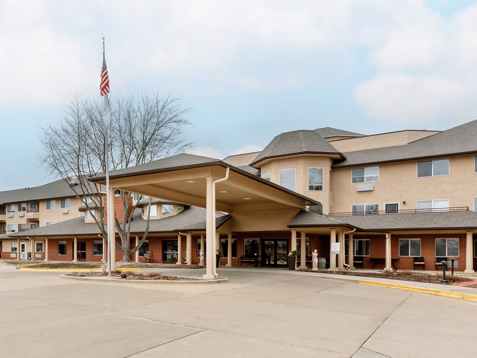 Front entrance of a multi-story senior living building with a covered drop-off canopy, an American flag, and a paved driveway.