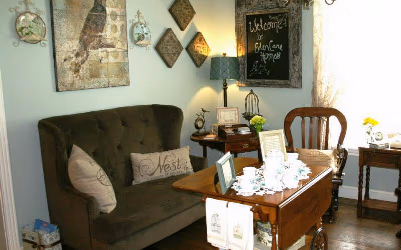 A cozy living room area with a dark green tufted loveseat adorned with two decorative pillows, a wooden side table with a lamp and framed pictures, a wooden chair, and a wooden tea cart set with a tea set and framed photos. The wall features decorative art pieces and a chalkboard sign that reads 'Welcome to Eden Care Homes'.