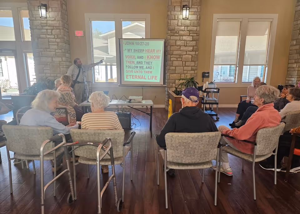 A group of elderly people seated in chairs facing a man who is pointing at a projection screen displaying a Bible verse from John 10:27-28. The room has large windows, stone walls, and wooden flooring, with some mobility aids visible.