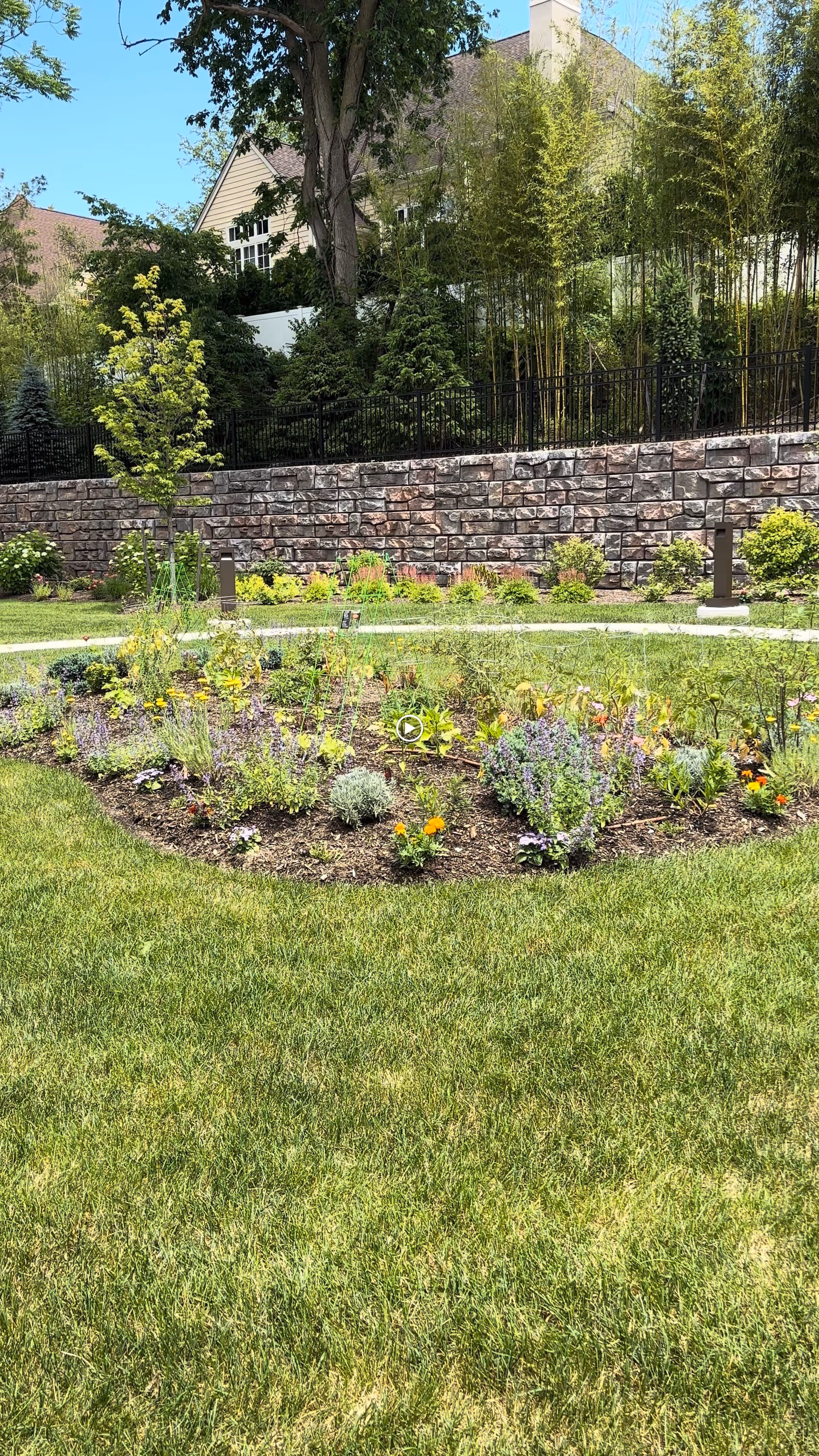 A landscaped garden area with a circular flower bed filled with various colorful flowers and plants. Surrounding the flower bed is a well-maintained green lawn. In the background, there is a stone retaining wall with a black metal fence above it, and behind the fence are tall trees and shrubs. A house with beige siding and a brown roof is partially visible behind the trees.