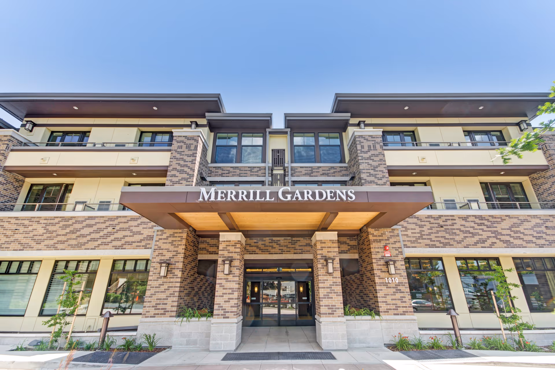 Front exterior view of Merrill Gardens senior living facility showing a modern three-story building with brick and beige paneling, large windows, and a covered entrance with the facility name displayed above.