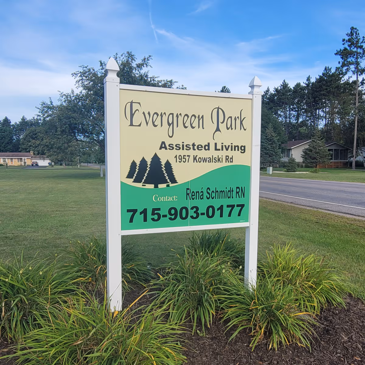 A sign for Evergreen Park Assisted Living Facility located at 1957 Kowalski Rd. The sign includes contact information for Rena Schmidt RN with the phone number 715-903-0177. The sign is set outdoors with grass, plants, trees, and houses visible in the background under a blue sky.