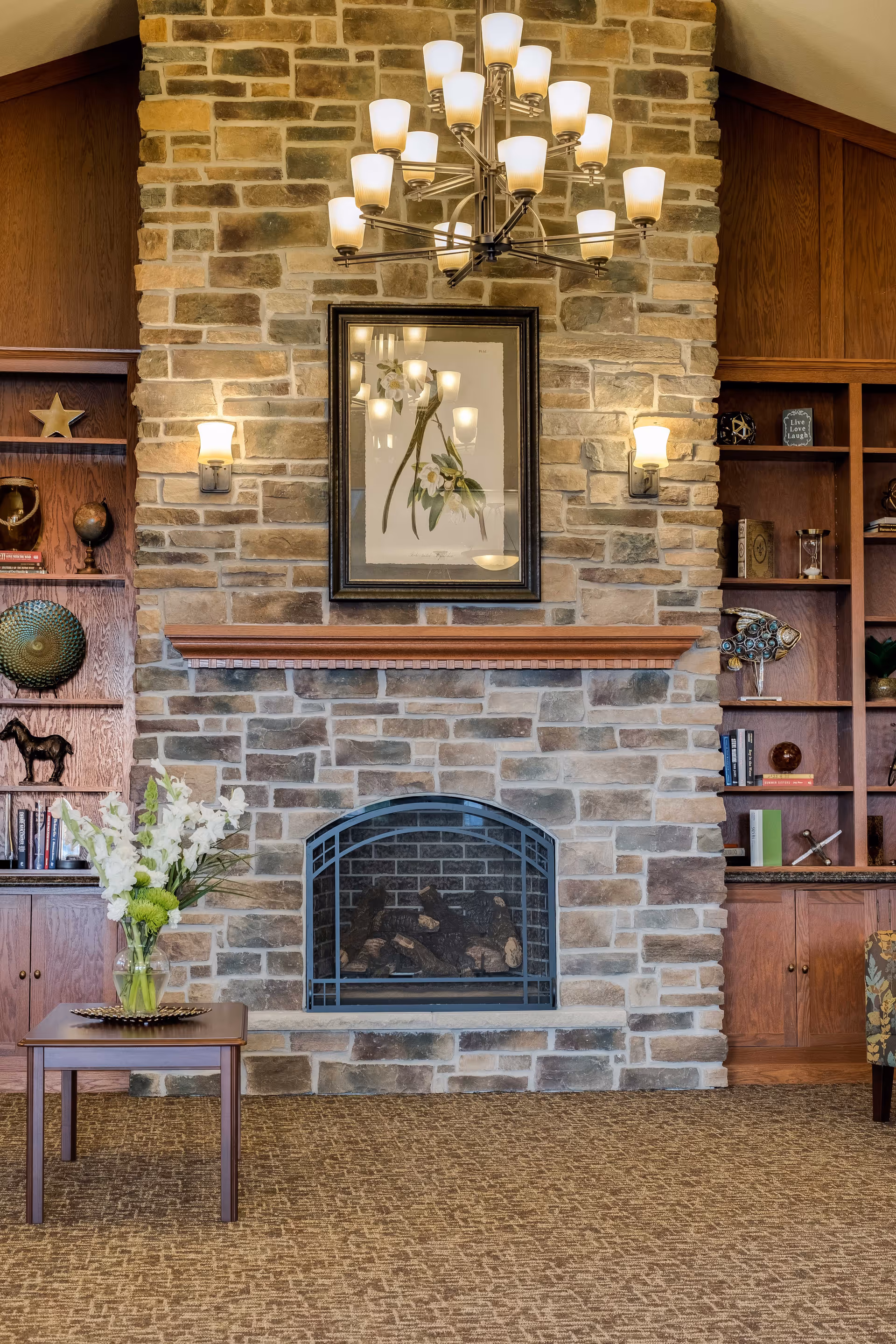 Cozy living room area with a stone fireplace in the center, flanked by wooden built-in shelves filled with decorative items and books. Above the fireplace is a framed floral artwork, and a modern chandelier with multiple lights hangs from the ceiling. A small wooden table with a vase of white flowers is placed in front of the fireplace.