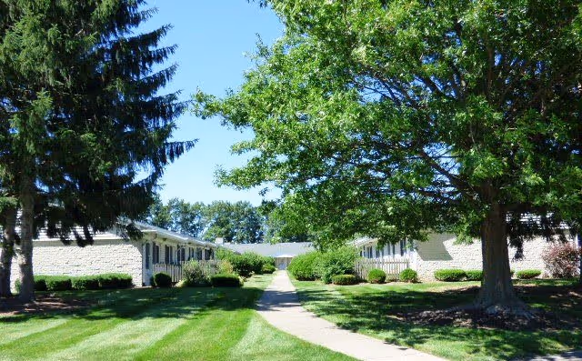 A sunny outdoor view of a retirement village with a paved walkway leading between two single-story buildings surrounded by well-maintained green lawns, bushes, and large trees under a clear blue sky.