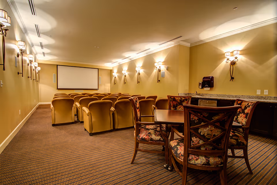 A cozy theater room with rows of beige armchairs facing a large blank projection screen. On the right side, there is a wooden table with four floral upholstered chairs. The walls are painted beige and decorated with multiple wall sconces emitting warm light. There is a countertop with a sink and a paper towel dispenser on the right side of the room.