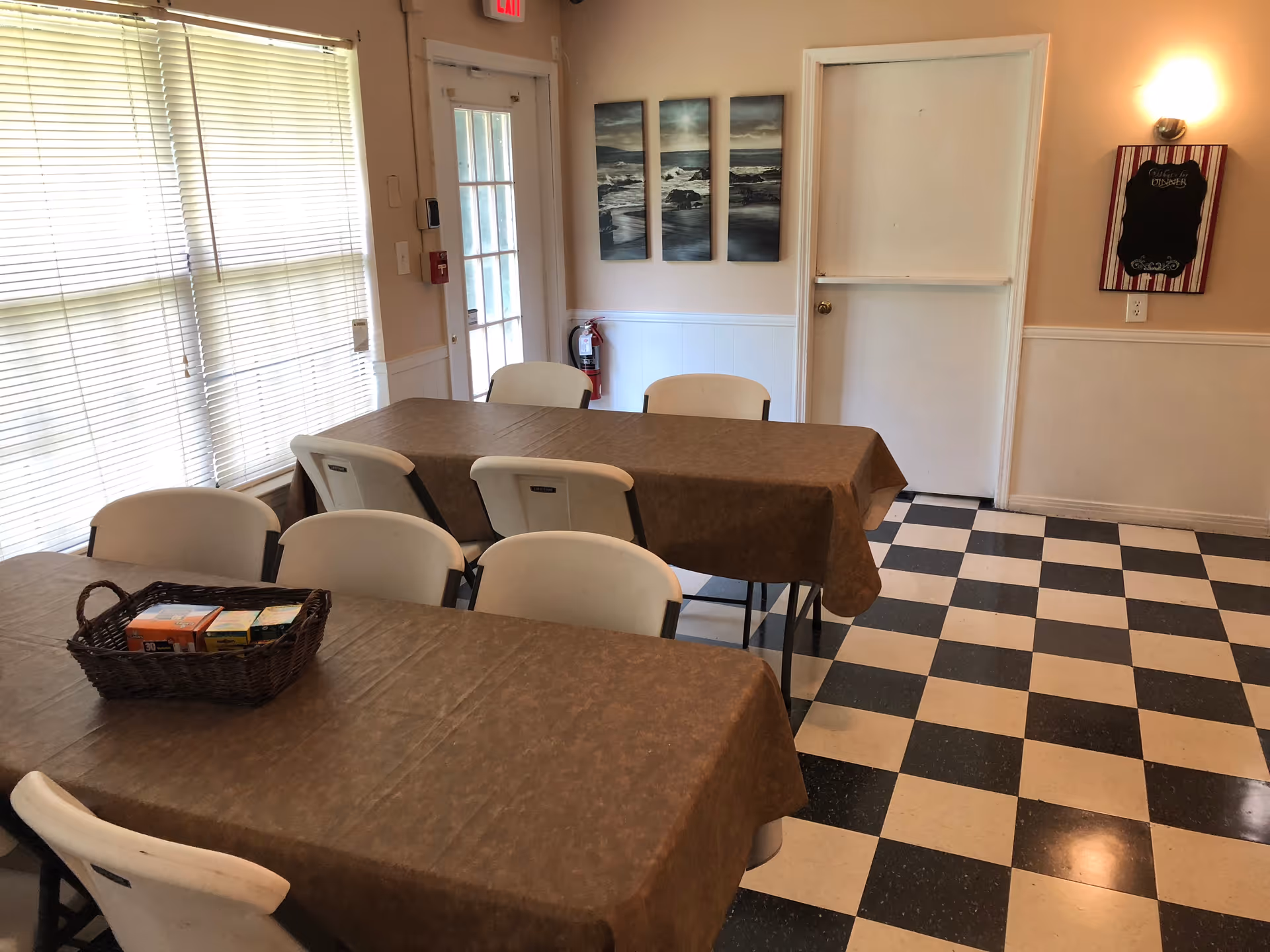 A dining area with two tables covered in brown tablecloths and surrounded by white chairs. A basket with various items is placed on the nearest table. The floor has a black and white checkered pattern. There is a door with a window and a fire extinguisher mounted on the wall next to it. Three framed pictures of ocean waves hang on the wall, and a small decorative board is mounted near another door.