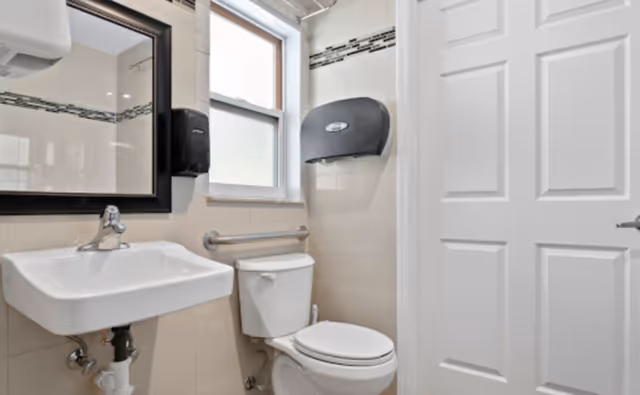 A clean and modern bathroom with a white sink, a toilet, a large mirror above the sink, a window, a handrail next to the toilet, and a wall-mounted paper towel dispenser.