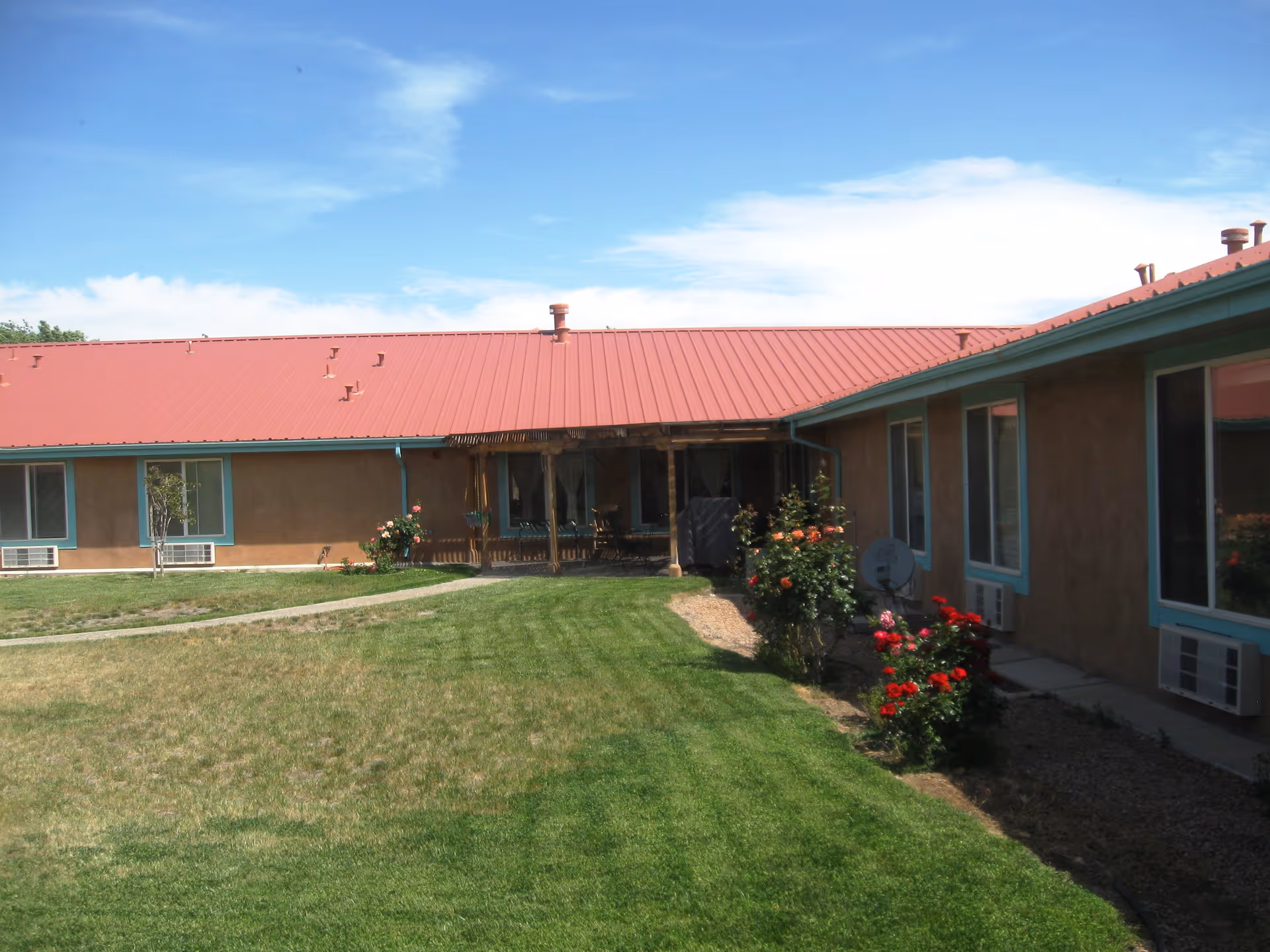 Single-story assisted living building with a red metal roof overlooking a green lawn, walkway, and flowerbeds in a courtyard.