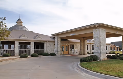 Exterior view of a single-story stone building with a covered entrance and a circular driveway, surrounded by neatly trimmed bushes and a lawn under a partly cloudy sky.