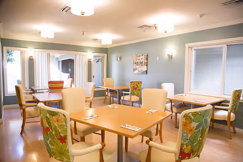 Bright communal dining room with multiple wooden tables and upholstered chairs, some with floral patterns, under ceiling lights.