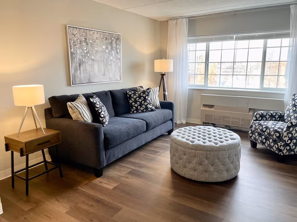 A cozy living room with a dark gray sofa adorned with patterned pillows, a round tufted ottoman in front, a patterned armchair to the right, and two lamps providing warm lighting. A large window with white curtains lets in natural light, and a framed abstract painting hangs on the wall above the sofa.