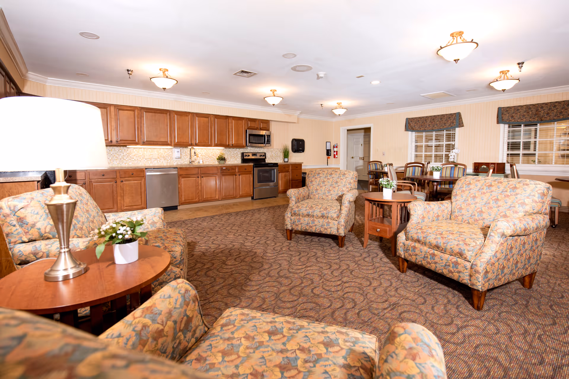 A spacious senior living common area featuring a kitchen with wooden cabinets, stainless steel appliances, and a mosaic tile backsplash. The room includes several upholstered armchairs with floral patterns arranged around wooden side tables with small potted plants. In the background, there is a dining area with multiple tables and chairs, windows with valances, and warm overhead lighting.