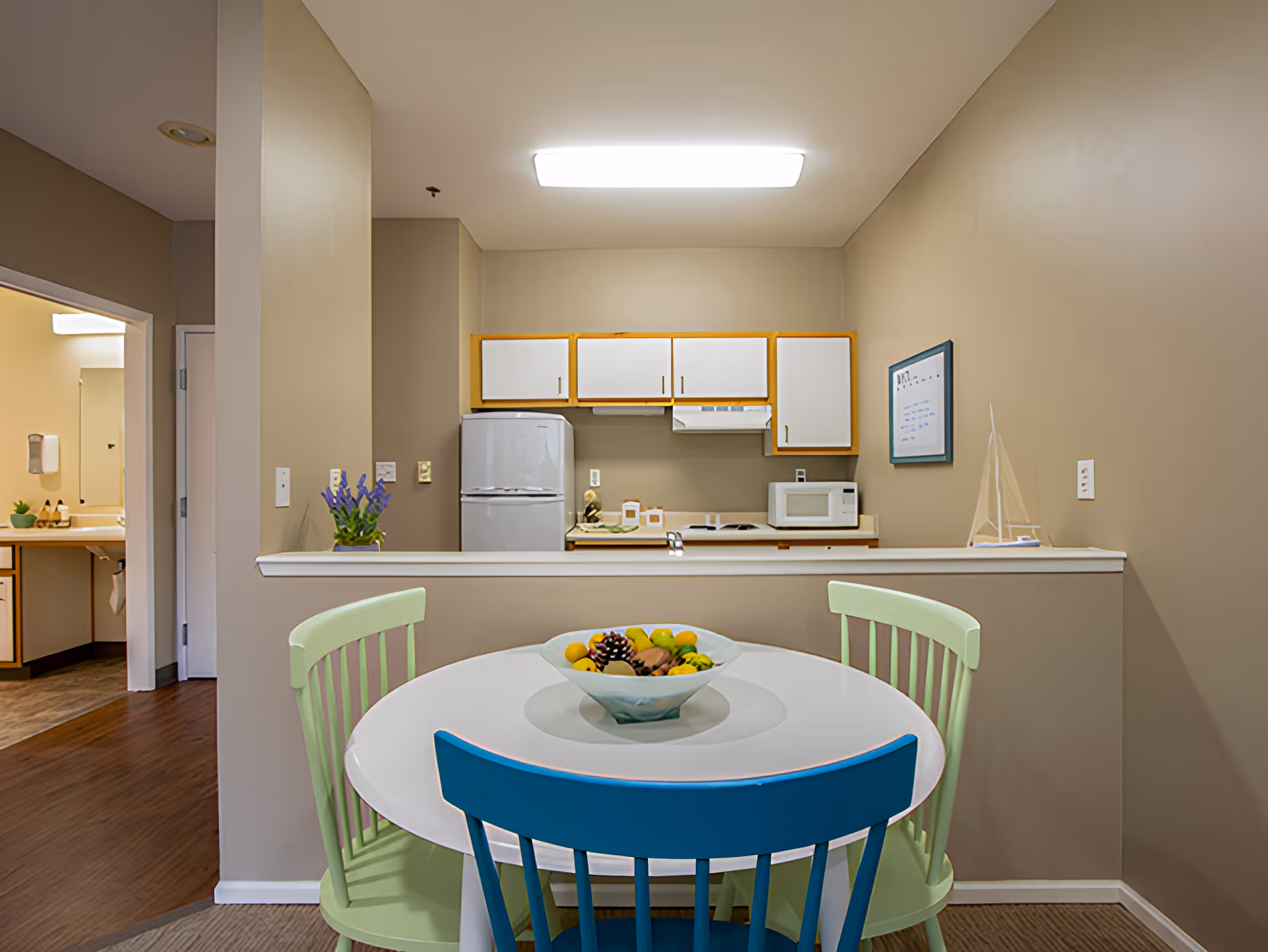 A small dining area with a round white table and three chairs, two light green and one blue. On the table is a decorative bowl filled with artificial fruit and pinecones. Behind the table is a half wall separating the dining area from a compact kitchen with white cabinets, a refrigerator, microwave, and stove. To the left, there is a doorway leading to a bathroom with a sink and mirror visible.