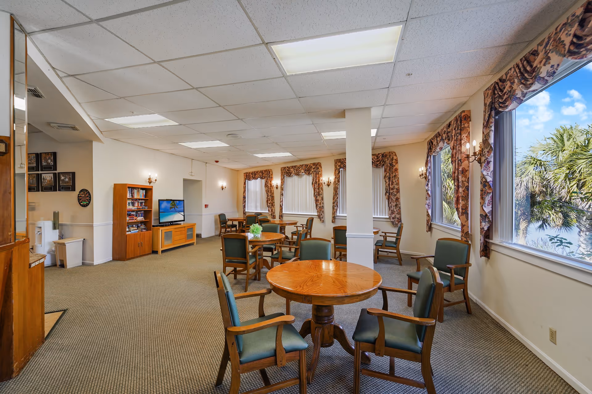 A well-lit common area in an assisted living facility with multiple round wooden tables surrounded by green cushioned chairs. The room has large windows with floral curtains, allowing natural light and a view of palm trees outside. There is a TV on a wooden stand and a bookshelf with books and games. The carpet is patterned, and the ceiling has fluorescent lighting panels.