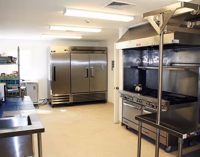 A clean commercial kitchen with stainless steel appliances including a large refrigerator, stove with oven, and a metal prep table. The room is well-lit with fluorescent lights and has a beige floor and white walls.