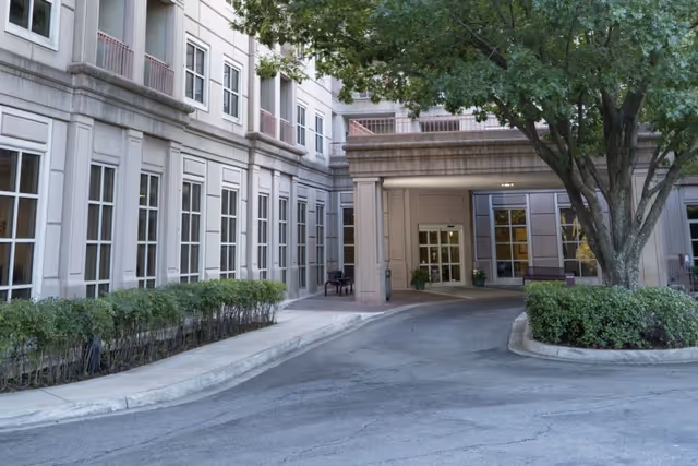 Entrance area of a healthcare facility with a covered drop-off zone, surrounded by a multi-story building with many windows, bushes lining the sidewalk, and a large tree with green leaves near the driveway.