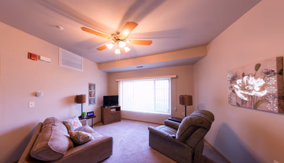 A warmly lit living room with a sofa, recliner, TV on a stand, lamps, ceiling fan, and a large window with vertical blinds.