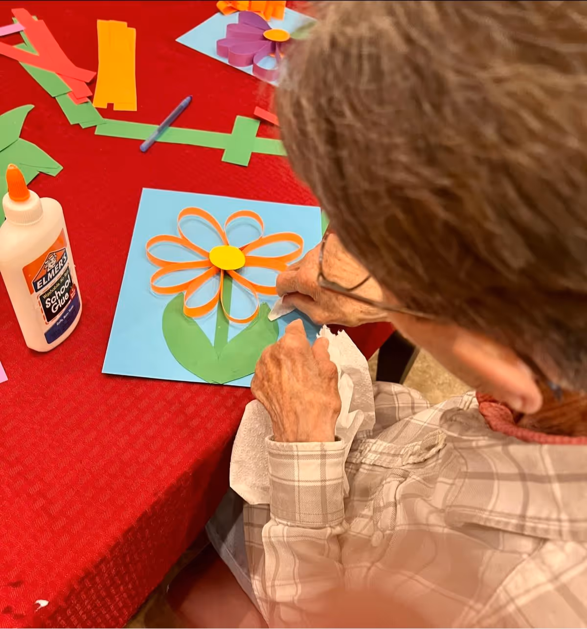 An elderly person wearing glasses and a plaid shirt is sitting at a red tablecloth-covered table, creating a paper flower craft using orange, yellow, and green paper strips glued onto a blue sheet. A bottle of Elmer's school glue is visible on the table.