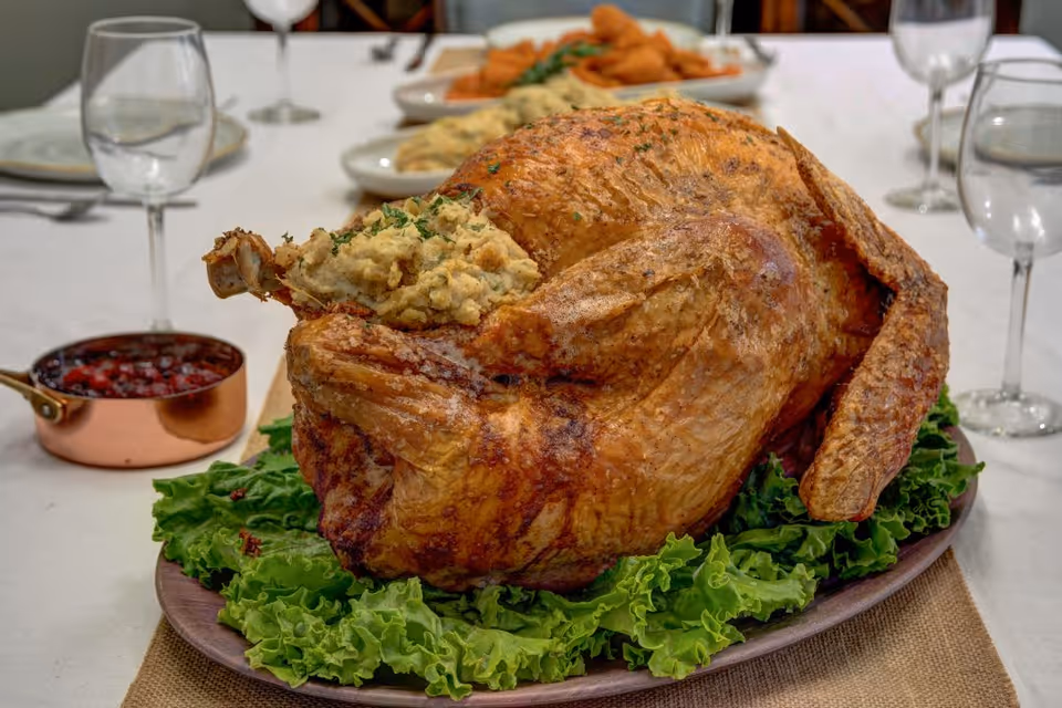 A roasted stuffed turkey placed on a wooden platter lined with green leafy lettuce, set on a white tablecloth with a small copper pot of cranberry sauce and empty wine glasses in the background.