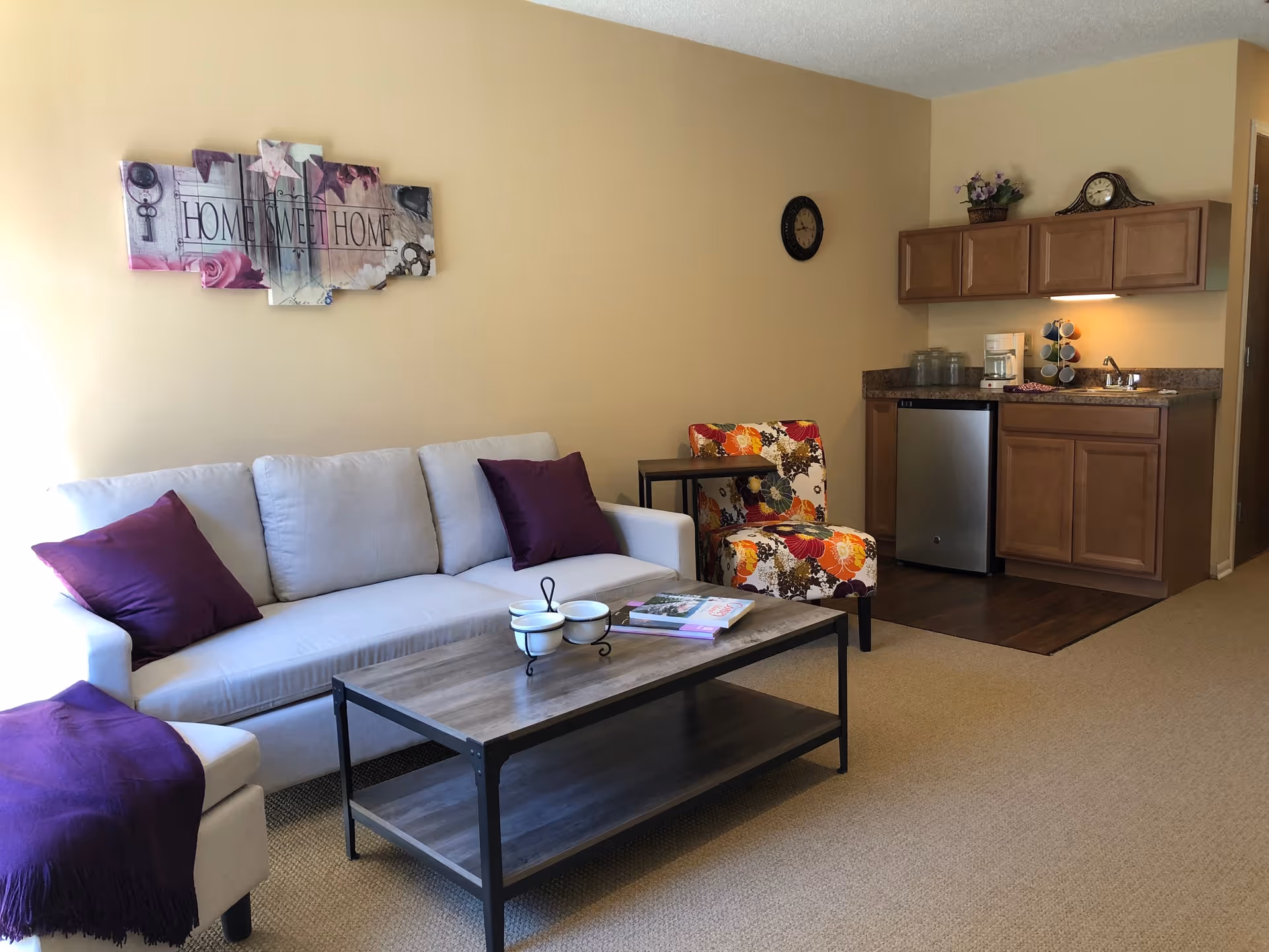 A cozy living room area with a light gray sofa adorned with two purple pillows and a purple throw blanket on the armrest. A wooden coffee table with a metal frame holds a decorative bowl and some magazines. Next to the sofa is a colorful floral-patterned armchair and a small side table. On the wall above the sofa is a decorative wall art piece that reads 'Home Sweet Home'. In the background, there is a small kitchenette with wooden cabinets, a mini fridge, a coffee maker, and a small sink. A clock and a small plant are placed on top of the cabinets.