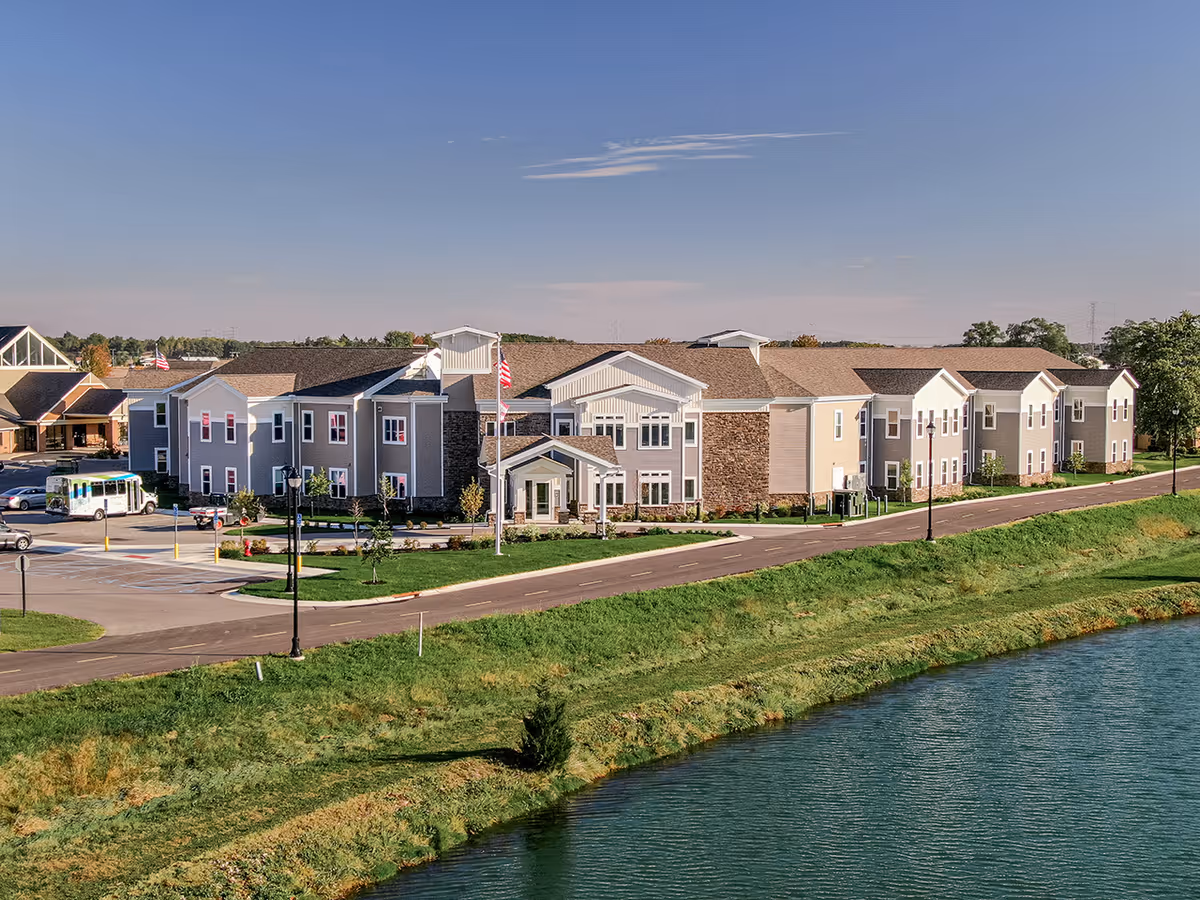Exterior view of Otterbein Pemberville SeniorLife Community, a large two-story building with beige and gray siding, multiple windows, and a main entrance with a covered porch. The building is situated next to a road and a body of water, with green grass and landscaping around it under a clear blue sky.