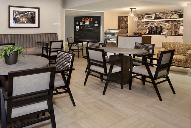 A cozy dining area with round tables and cushioned chairs arranged neatly. There is a built-in bench along one wall with a framed black and white photograph above it. In the background, a small counter area with coffee dispensers and a display case is visible, along with a blackboard sign that reads 'ZEST Takeout to go available!'. The room has light-colored flooring and neutral-toned walls with decorative stone accents.
