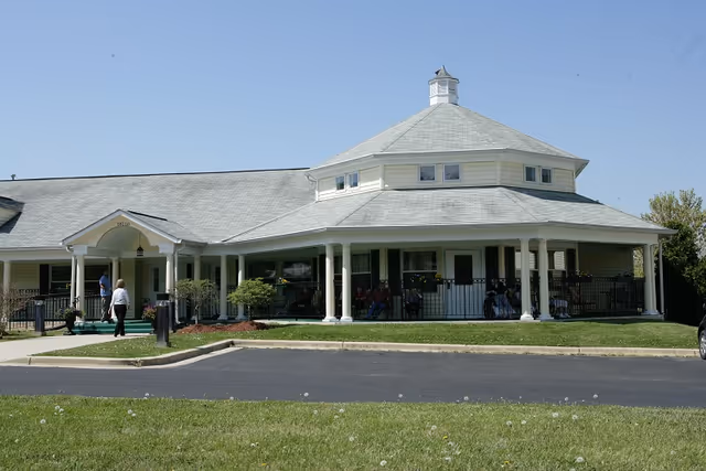 Front exterior of a single-story senior living facility with a wraparound porch and a few people near the entrance.