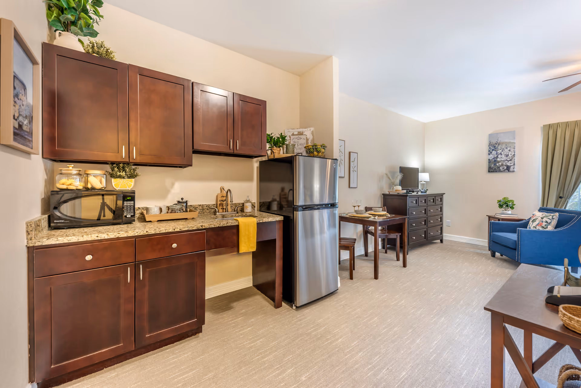 A bright and cozy interior space featuring a small kitchen area with dark wooden cabinets, a granite countertop, a microwave, and a stainless steel refrigerator. Adjacent to the kitchen is a dining area with a wooden table set for two, and a living area with a blue armchair, a dark wooden dresser with a TV on top, and a side table with a plant. The room has beige walls, carpeted flooring, and natural light coming through a window with green curtains.