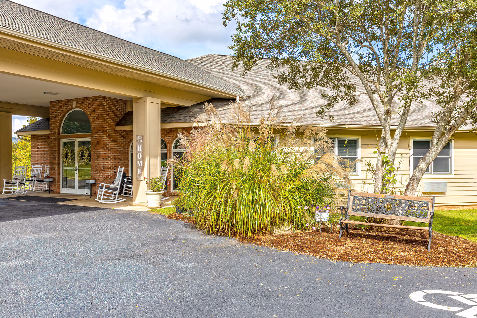 Entrance of a senior living facility with a covered driveway, brick and beige siding exterior, rocking chairs on the porch, a bench near a tree, and ornamental grasses in a landscaped area.