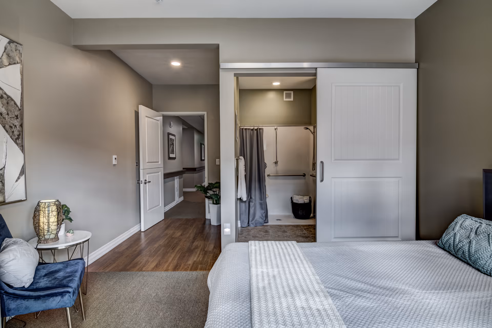 A bedroom in an assisted living facility featuring a bed with a white textured blanket and a blue decorative pillow. To the left is a small round table with a lamp and a plant, next to a blue upholstered chair. The room has beige walls and a mix of carpet and wood flooring. A sliding door reveals an accessible bathroom with a shower equipped with grab bars and a gray shower curtain. An open door leads to a hallway with framed pictures and plants.