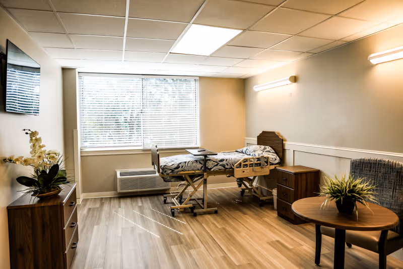 Private senior living bedroom with an adjustable hospital-style bed, bedside furniture, TV, and seating by a large window.