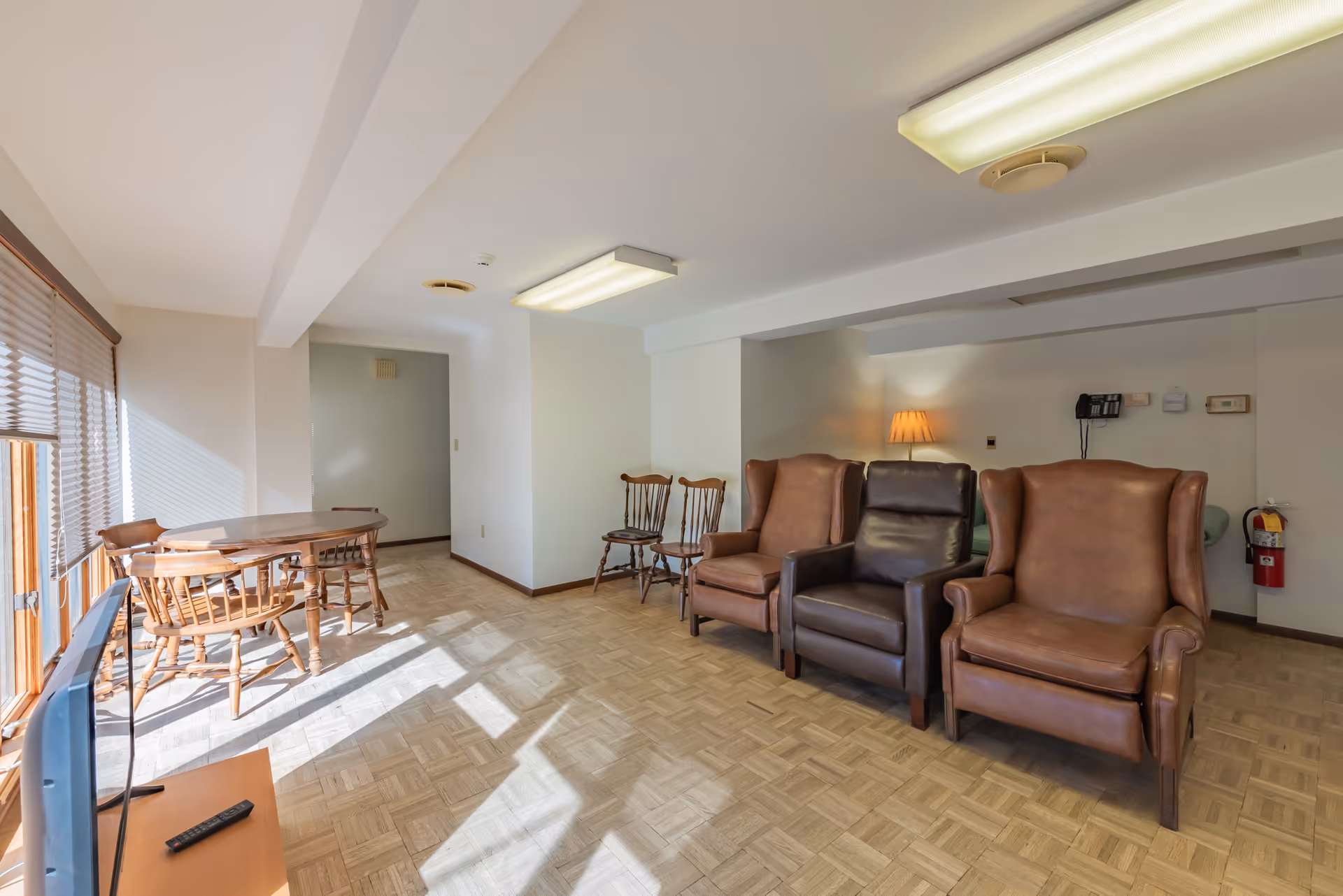 A bright living room area with large windows on the left letting in sunlight. The room features a wooden dining table with chairs near the windows, three armchairs including two brown leather wingback chairs and one dark brown leather recliner, additional wooden chairs against the wall, a floor lamp, and a fire extinguisher mounted on the wall.