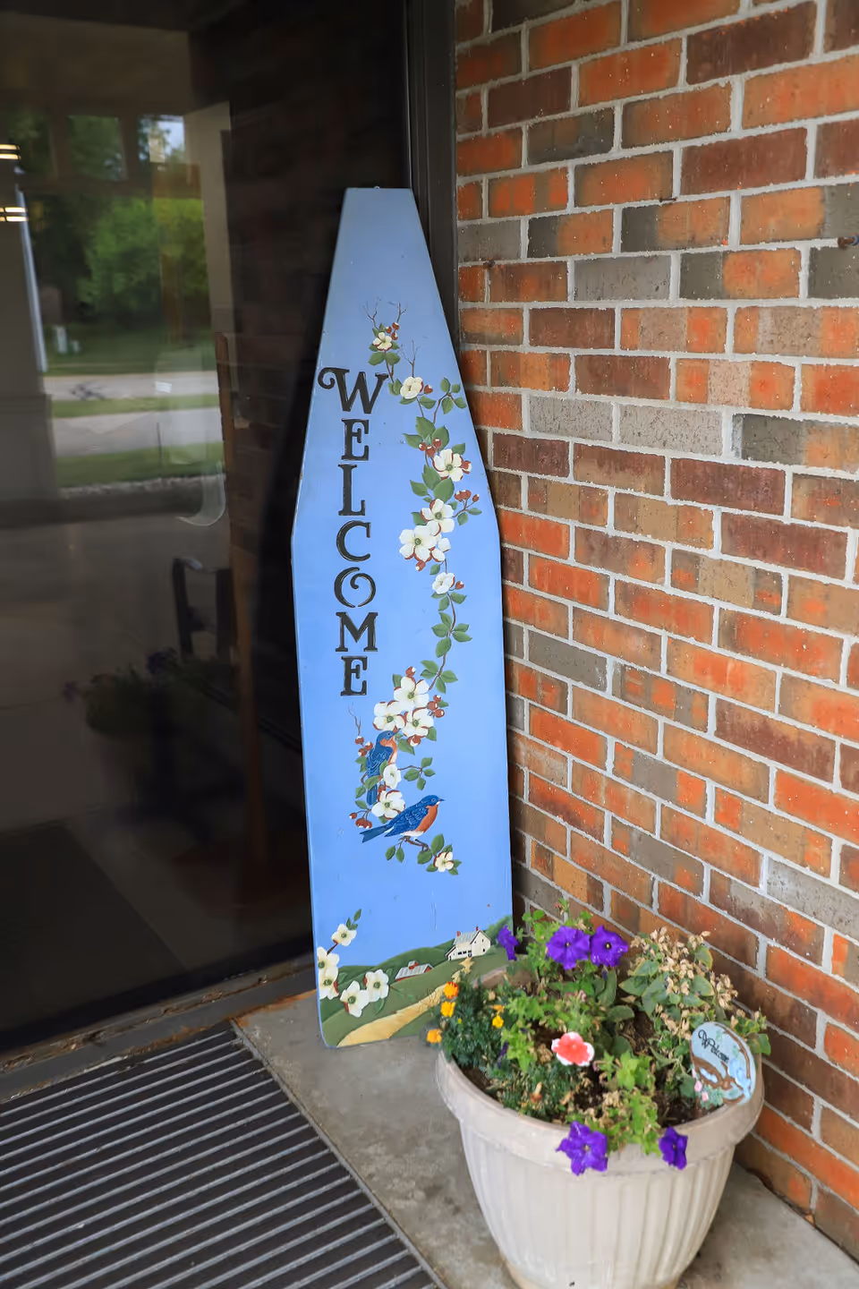 A blue wooden welcome sign decorated with painted white flowers and two birds leaning against a brick wall near a glass door entrance. Next to the sign is a white planter pot containing various colorful flowers including purple and pink blooms.