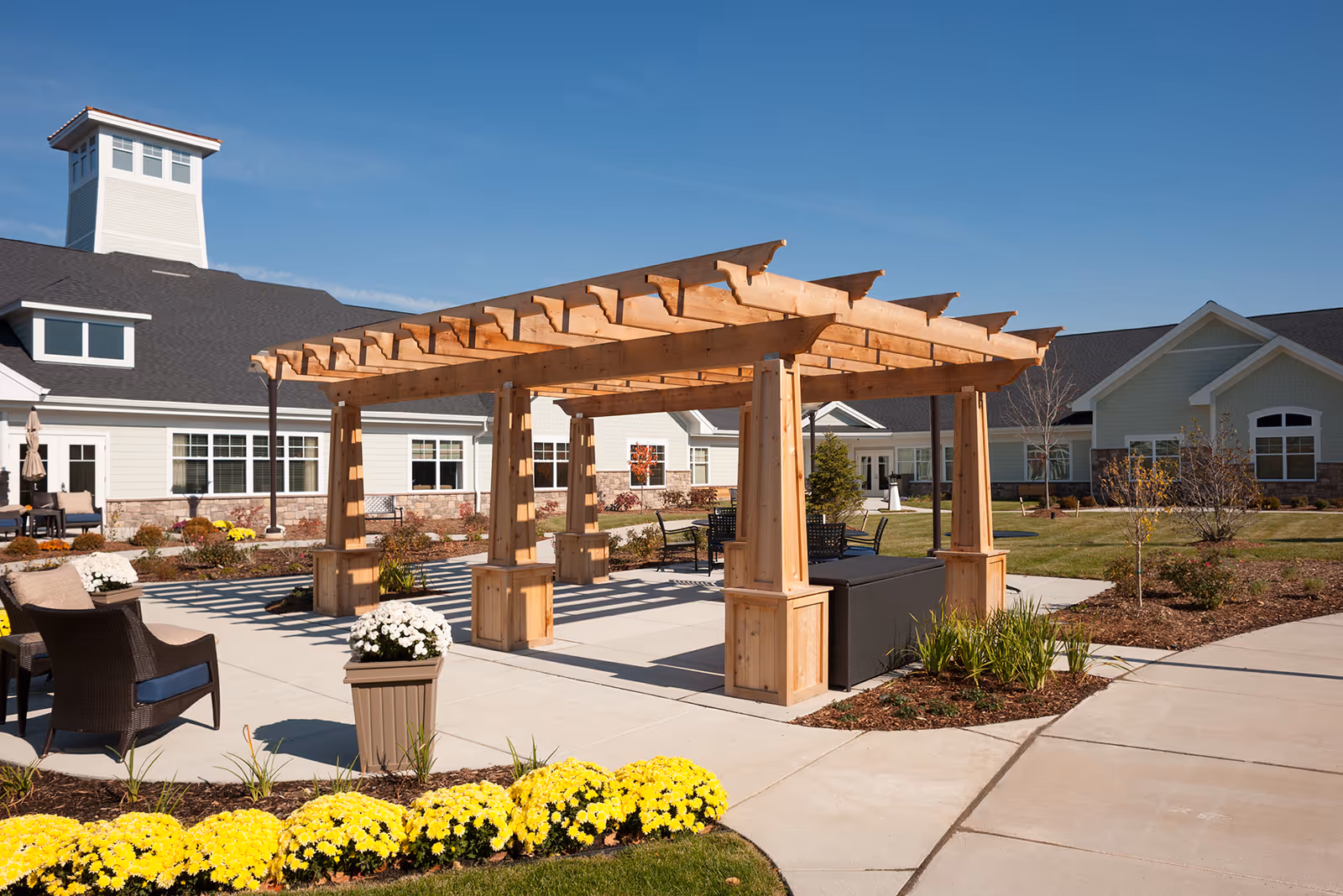 Outdoor patio area at Pomeroy Senior Living of Northville featuring a wooden pergola with seating underneath, surrounded by landscaped garden beds with yellow flowers and shrubs, and the senior living facility building in the background under a clear blue sky.