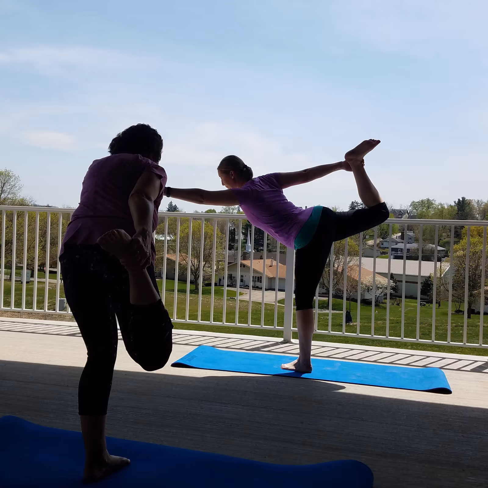 Two people practicing yoga on blue mats on a balcony with a white railing, overlooking a green landscape with trees and houses under a clear sky.
