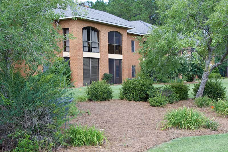 Exterior view of a two-story brick building surrounded by green trees, bushes, and landscaped garden beds with mulch and grass.